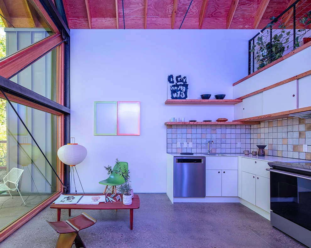 Interior of a kitchen with floating wooden shelves, tiled backsplash, and stainless steel appliances next to floor-to-ceiling glazing.