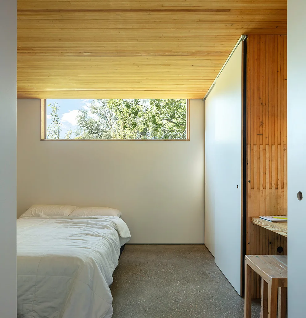A minimalist bedroom features a horizontal window, wooden ceiling, and a concrete floor.