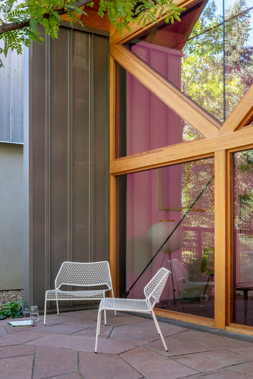 The patio area of the A.D.U. project features white metal mesh chairs, stone paver flooring, and large triangular wooden framed windows with tinted glass.