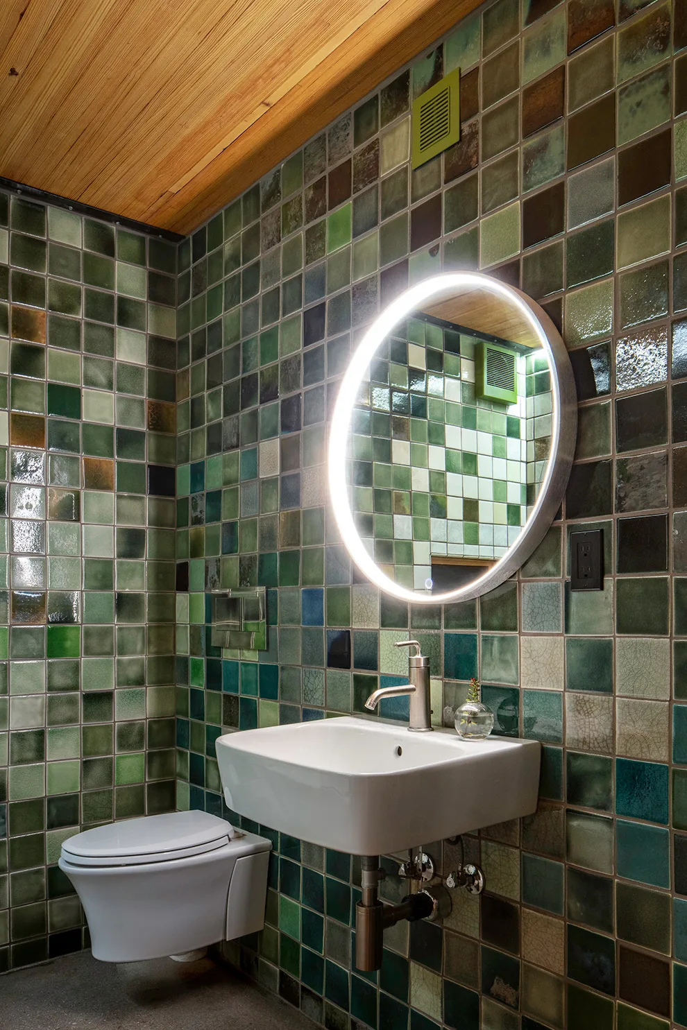 Bathroom with green square glazed tiles, wall-mounted toilet, sink, and a round illuminated mirror.