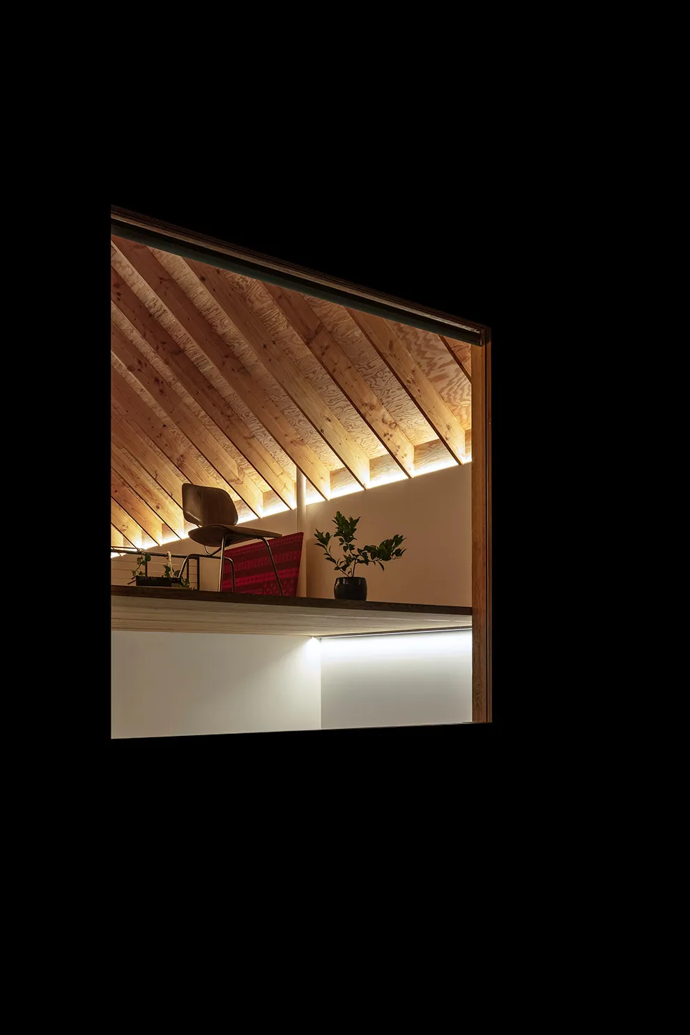 Room interior features exposed angled wooden beams and simple furnishings with plants on a shelf.
