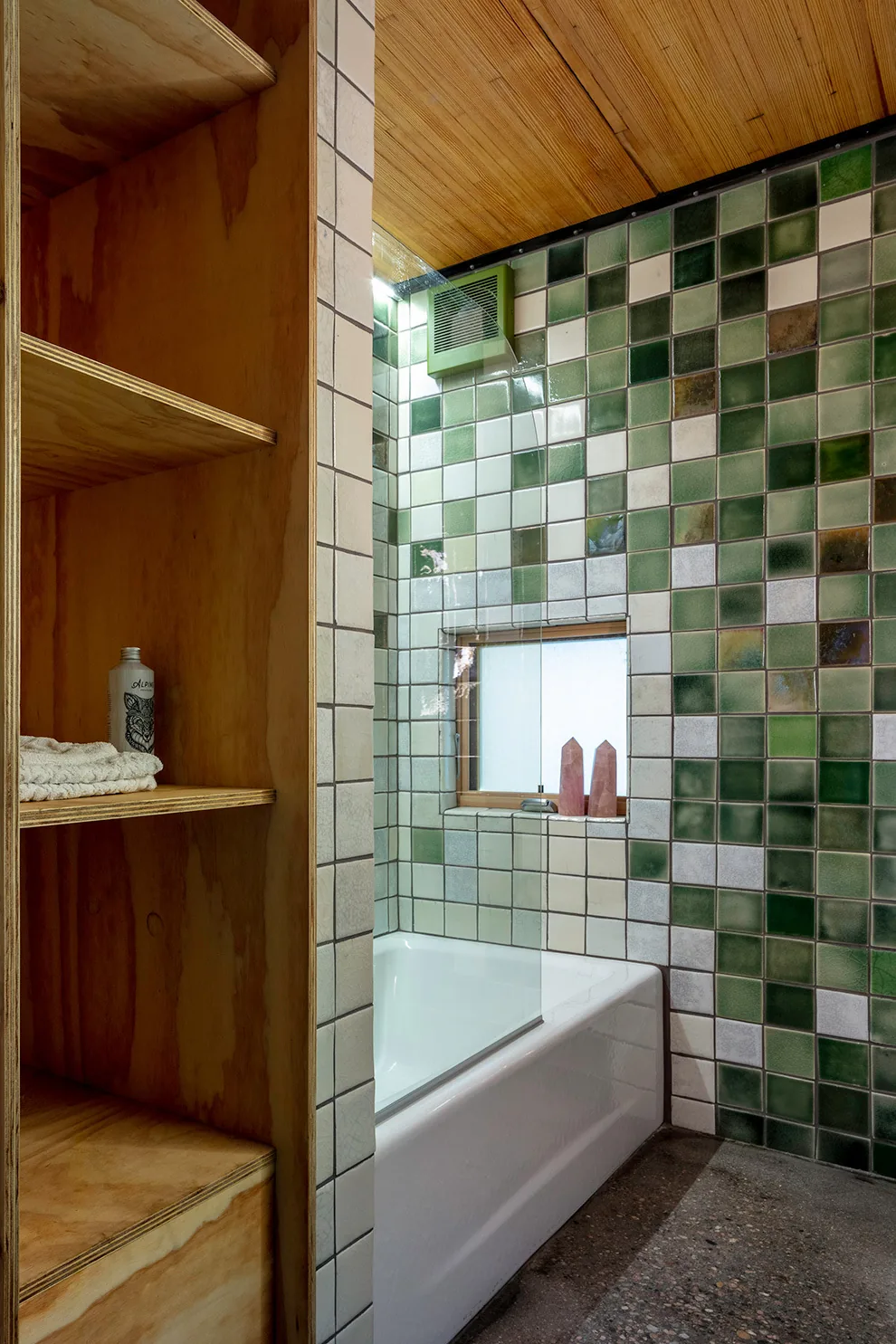 A bathroom features a white bathtub surrounded by green and white square tiles, a small window, and wooden shelving.