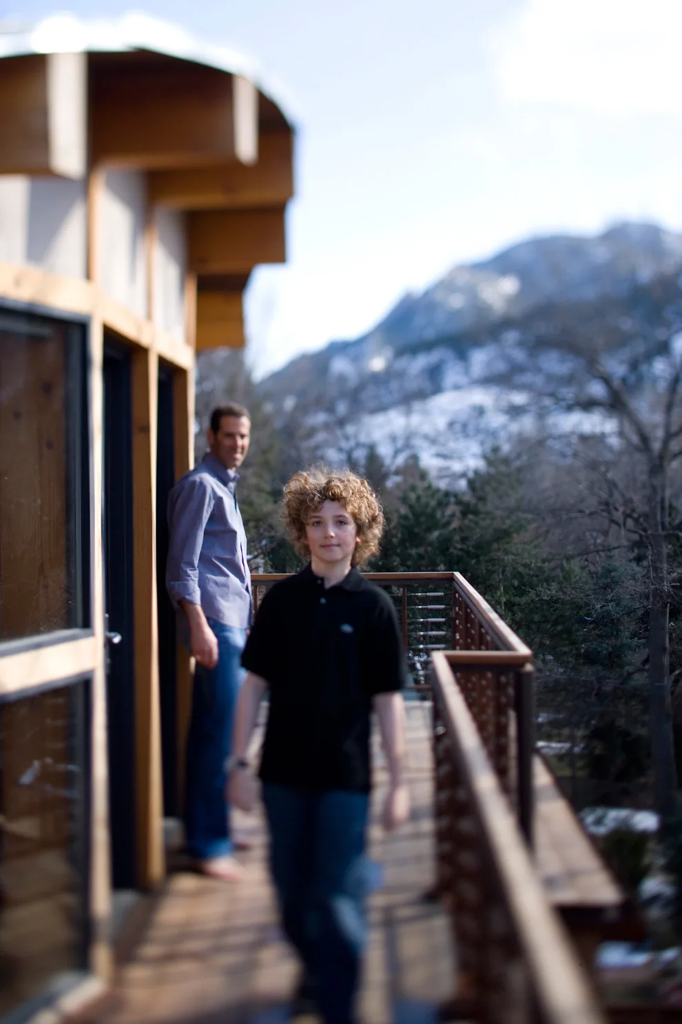 Wood balcony with railing attached to a building with large windows, with mountain landscape in the background.