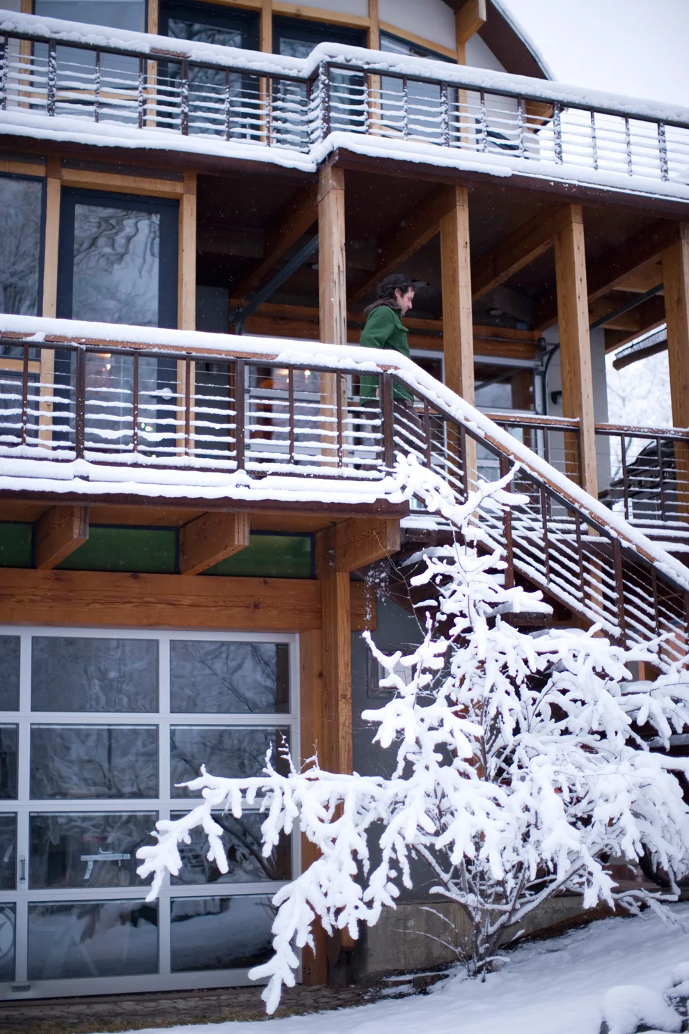 Wood and metal exterior stair with snow on railings on a two-story house with extensive glazing and a garage door.