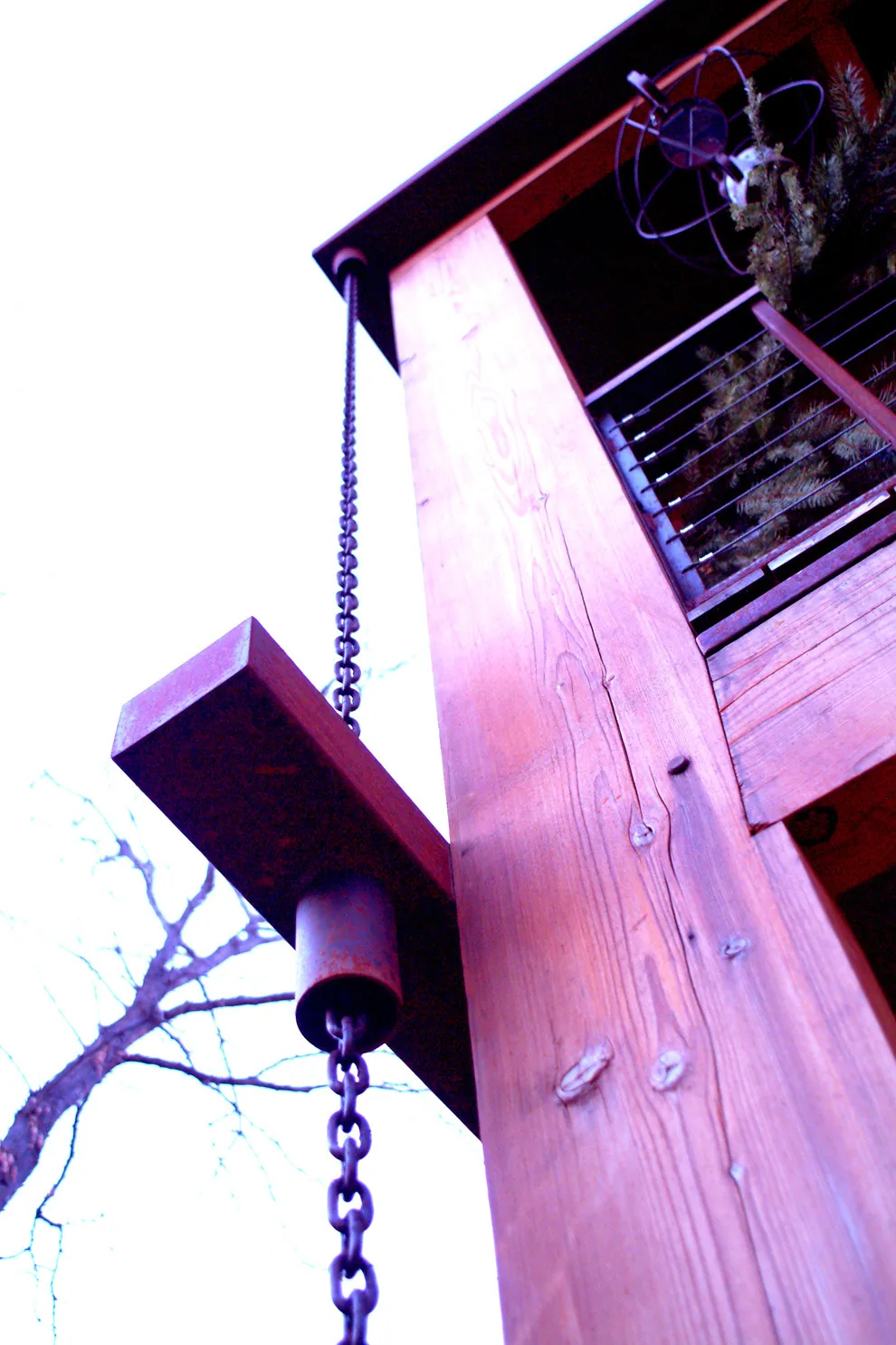 A close-up detail of a wooden support post with a hanging metal chain next to a balcony guarded by cable railing.