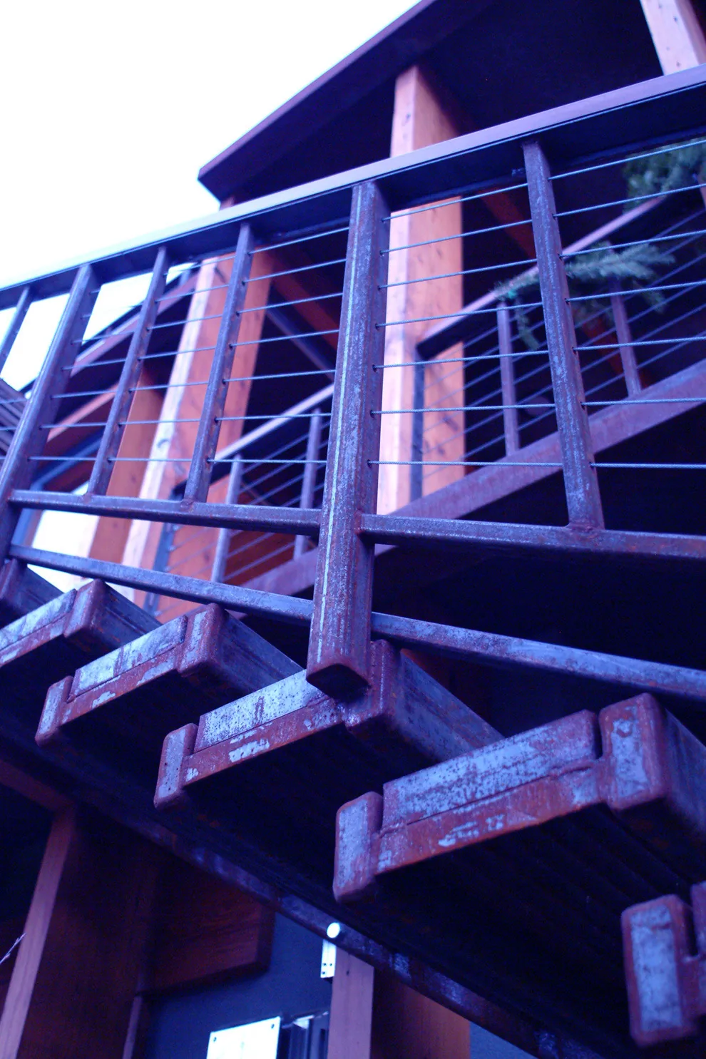 Rust-stained metal staircase and cable railing detail on exterior balcony with wood structural elements.
