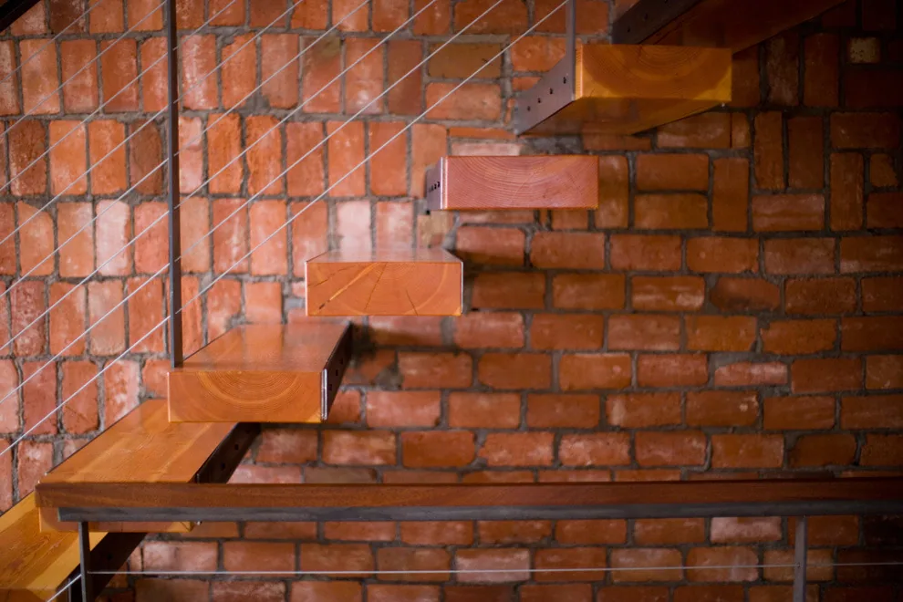 A close-up view of floating wooden stairs attached to an exposed brick interior wall with metal cable railing.