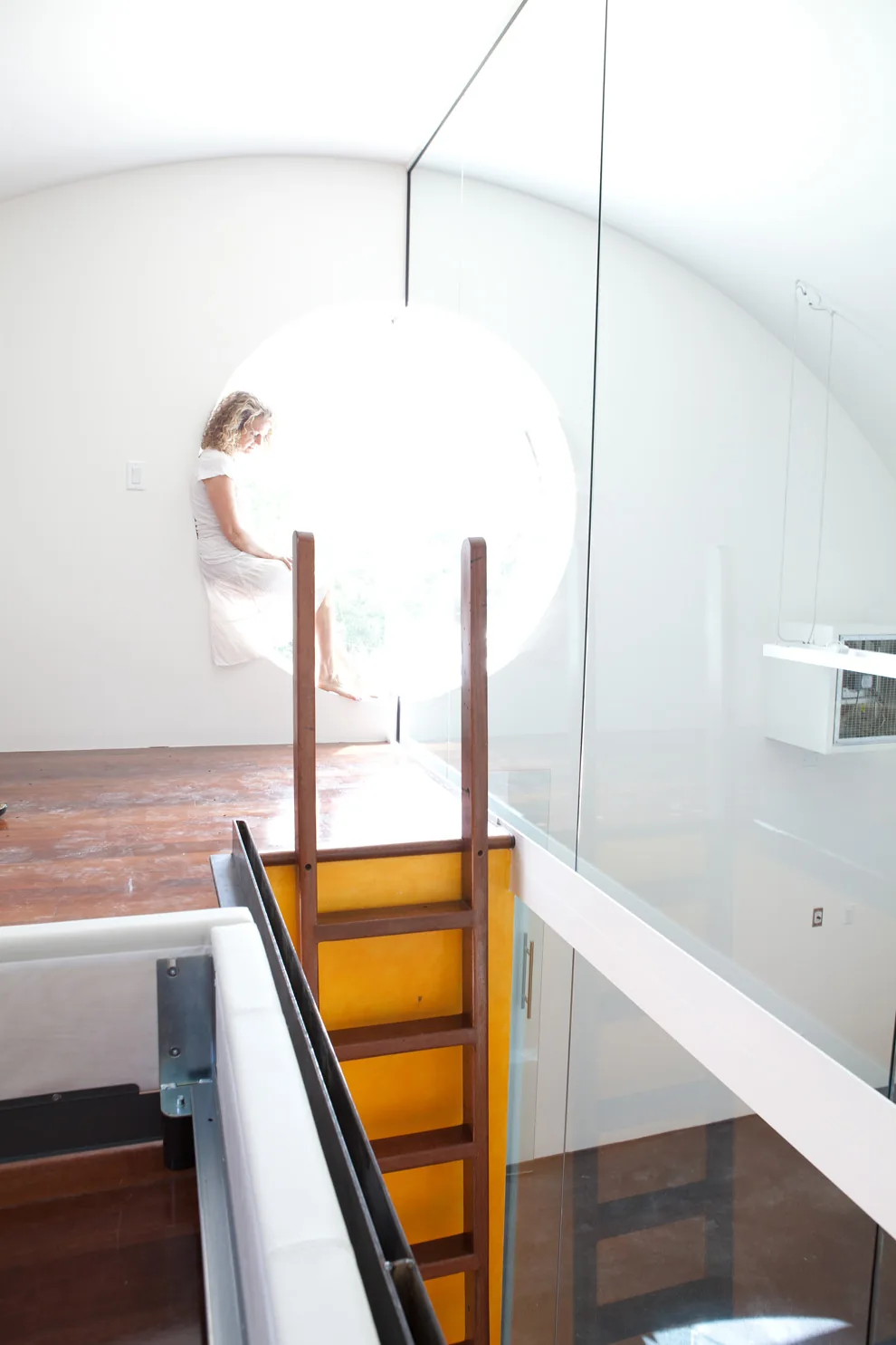A woman sits in a circular window seat near a wooden ladder inside a modern interior with glass railing.