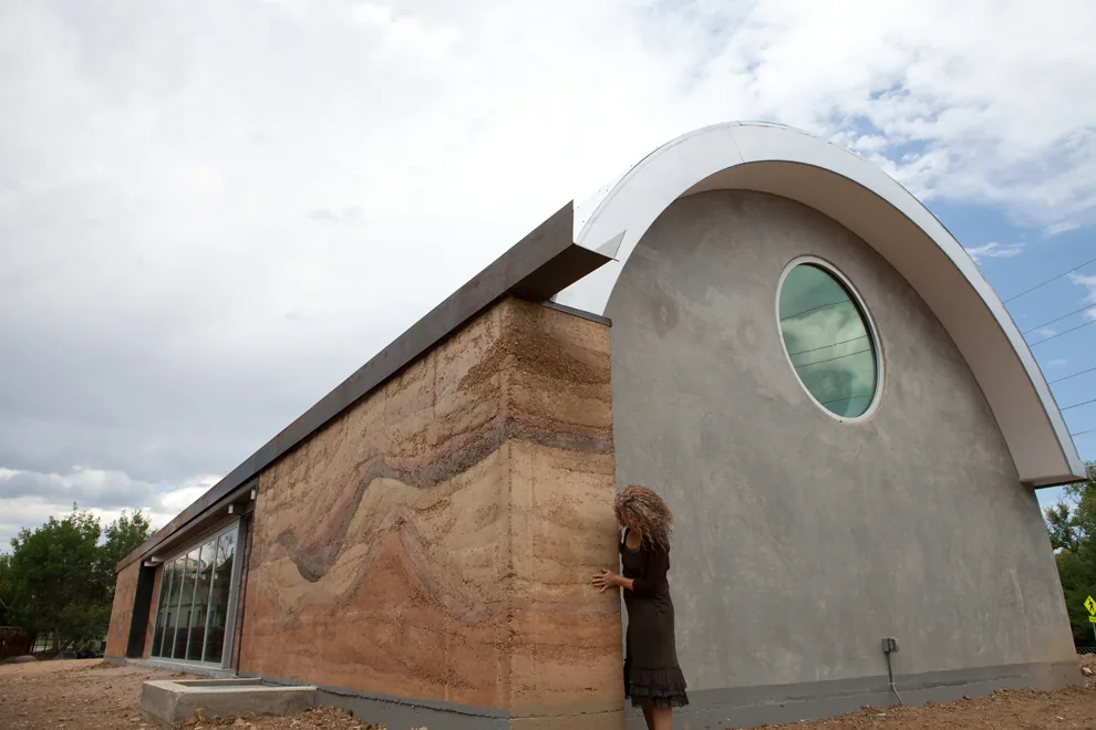 Exterior view of a building with a curved roof, rammed earth wall, and a circular window on the plastered wall.