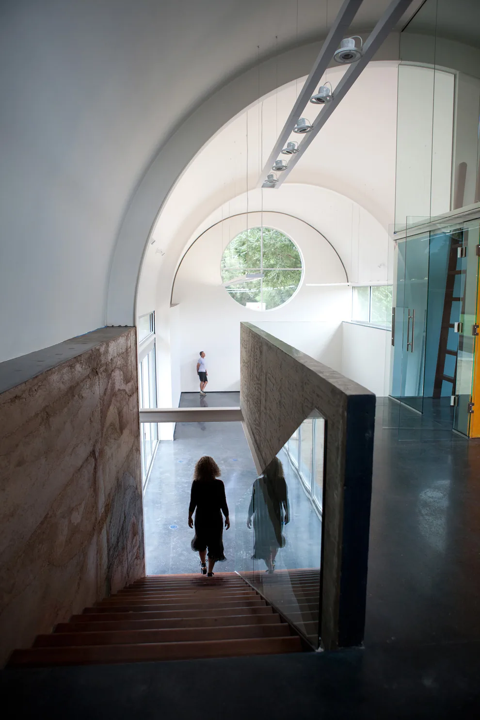 Interior space features vaulted ceilings, a large circular window, concrete stairs with glass railing, and polished concrete floors.