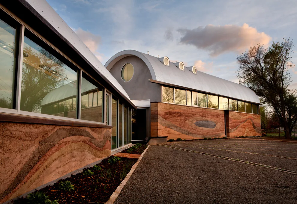 Exterior view of a building with curved metal roof and large windows above rammed earth walls at sunset.