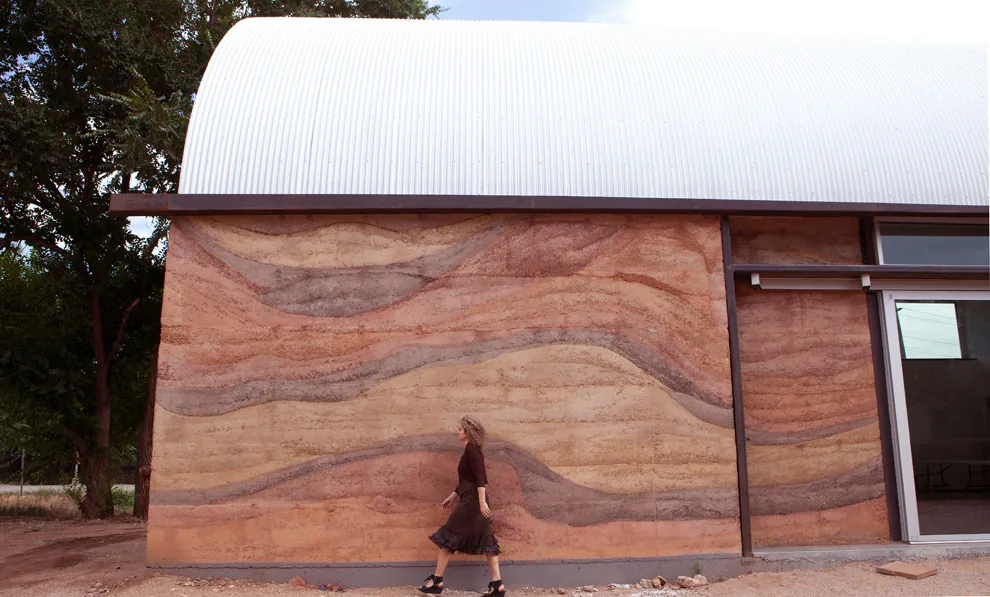 Building side with rammed earth wall under a curved metal roof, featuring a sliding glass door.
