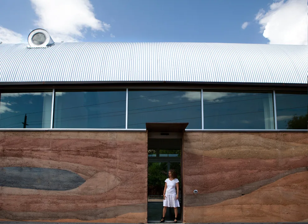 The building has a corrugated metal curved roof with clerestory windows and rammed earth walls with a single entrance where a person stands.