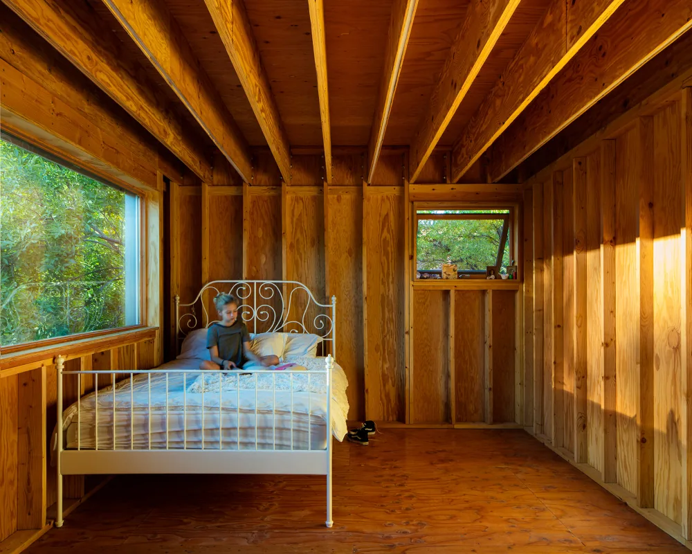 Bedroom with exposed wooden framing, visible ceiling joists, and natural light from large window and smaller casement window.