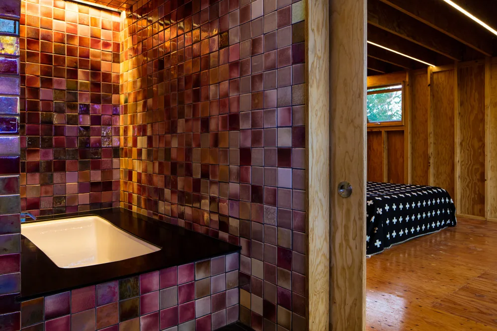 Bathroom sink area with multicolored square tile walls next to a wood-paneled bedroom with a patterned bedspread.