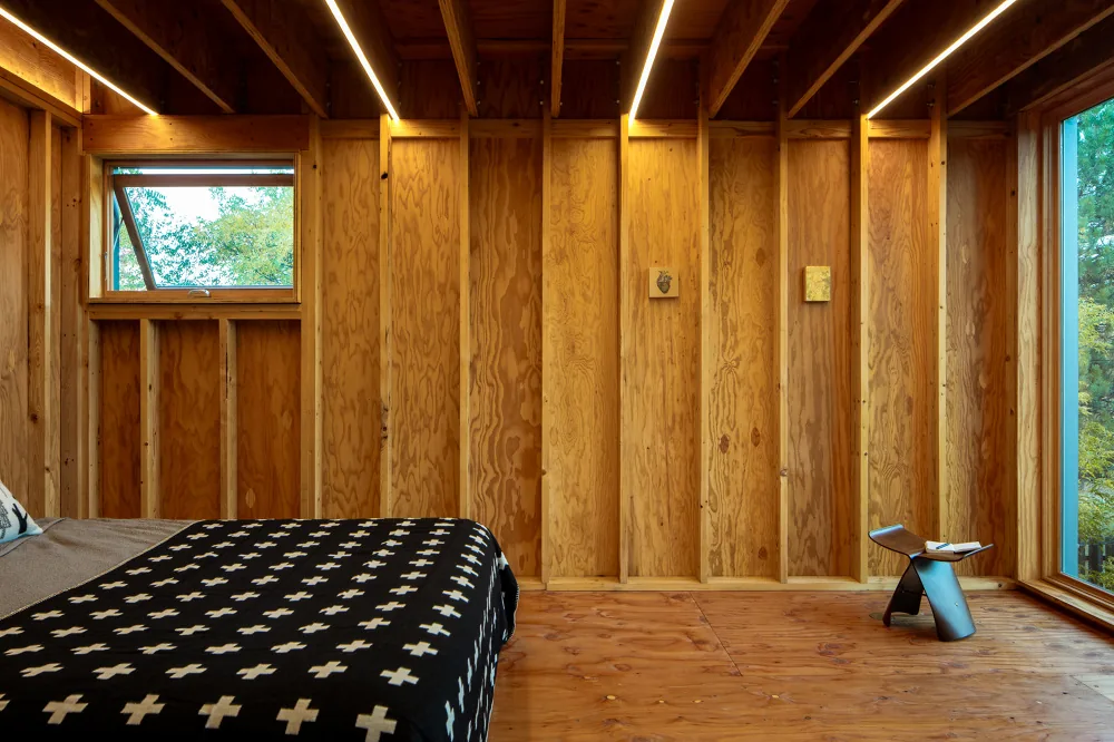 Bedroom interior showing exposed wood studs and beams with natural light from two windows.