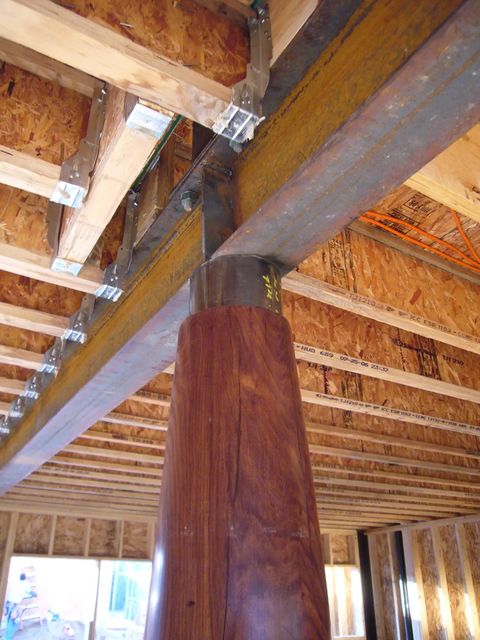A wooden post with a metal mounting base supports a large steel beam in a wood-framed building under construction.