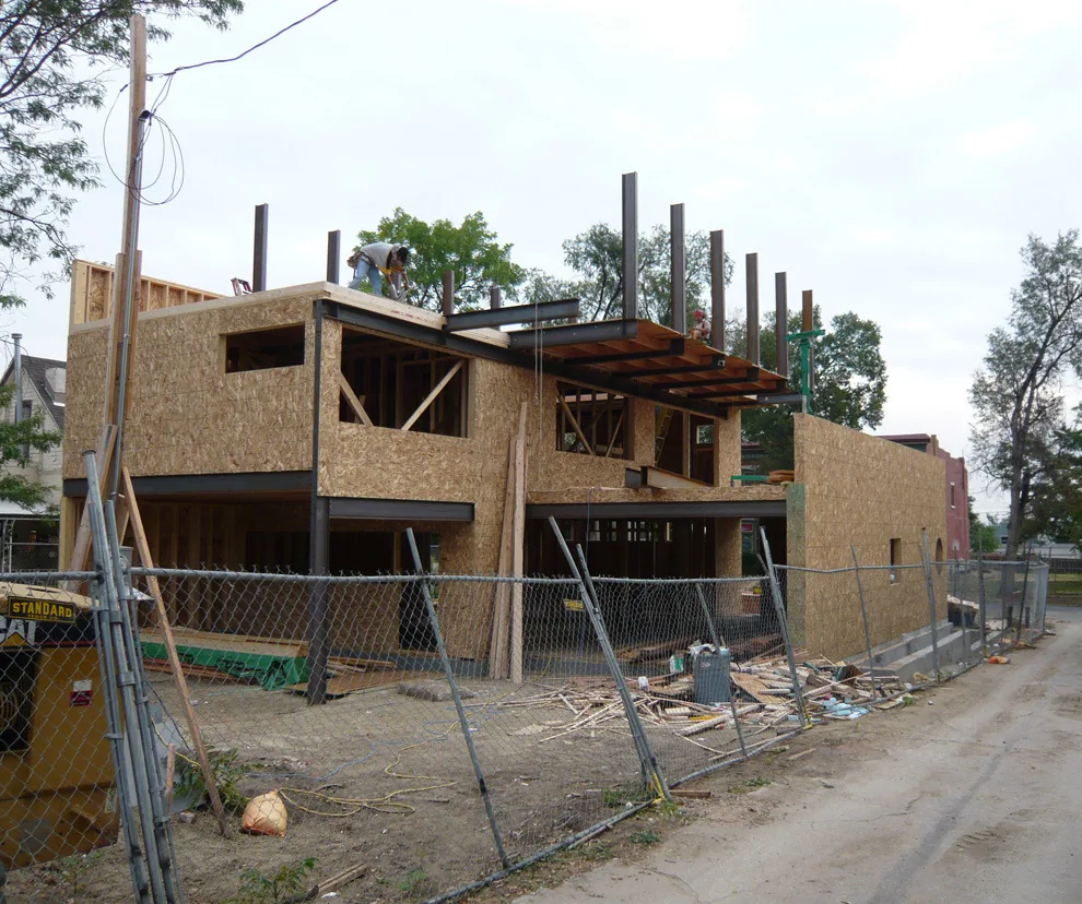 Construction phase of a two-story building showing steel framing and OSB sheathing on site fenced with metal chain link.