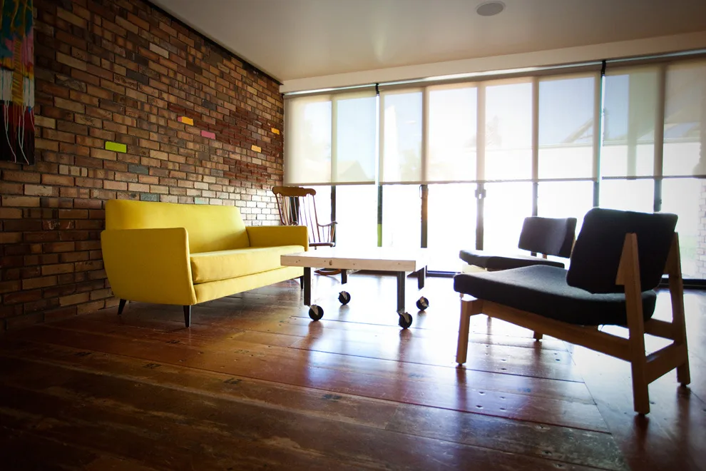 Living room features exposed brick wall, large windows with roller shades, and a mix of seating furniture around a wood and metal coffee table with casters.