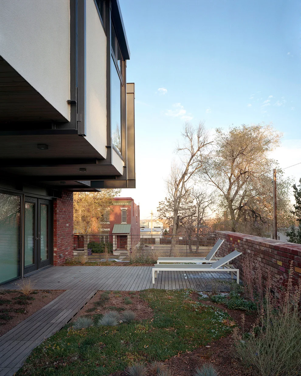 Modern house features a cantilevered upper floor with large windows, a wooden deck, and a brick garden wall with lounge chairs.