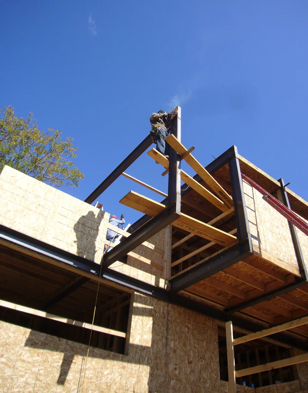 A construction worker welds the steel frame of a building while oriented strand board sheathing is installed on lower walls.