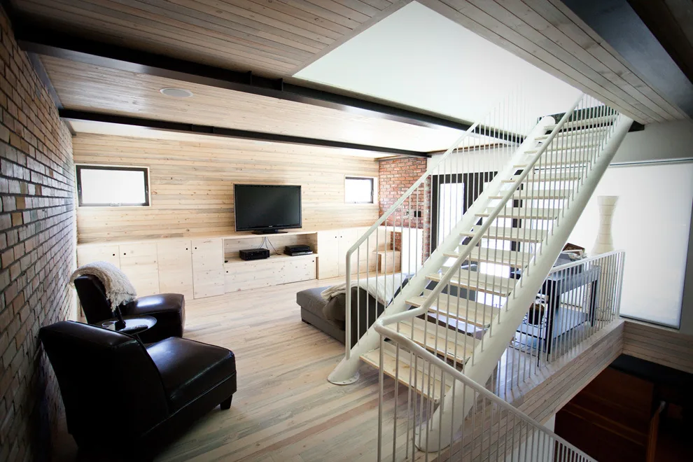 Living room with wood paneling, exposed brick walls, and a white metal open-riser staircase.