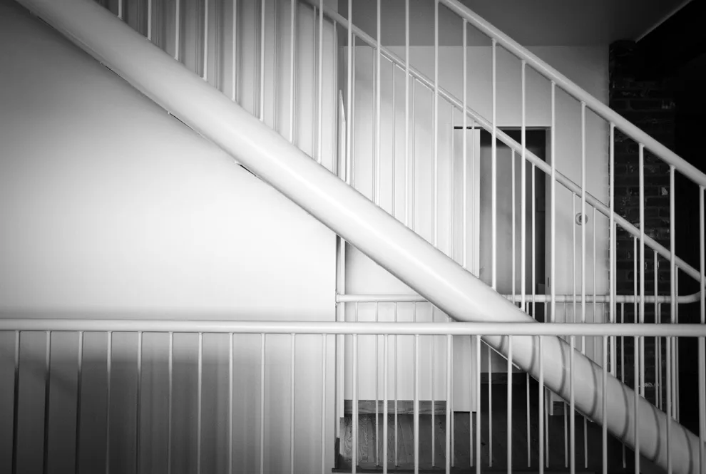 Interior black and white photograph focusing on a metal staircase with vertical balusters and a diagonal handrail.