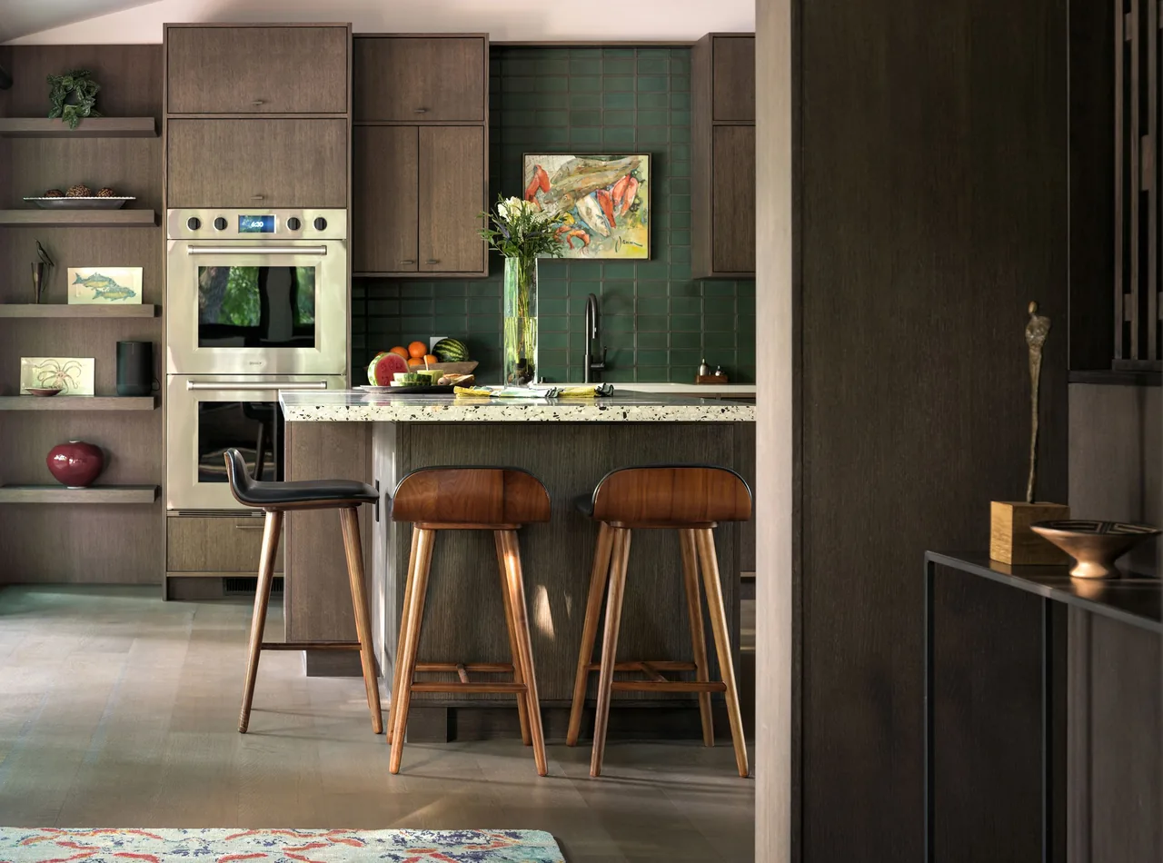 Modern kitchen features dark wood cabinetry, terrazzo island countertop, green tile backsplash, and built-in double oven with bar seating.