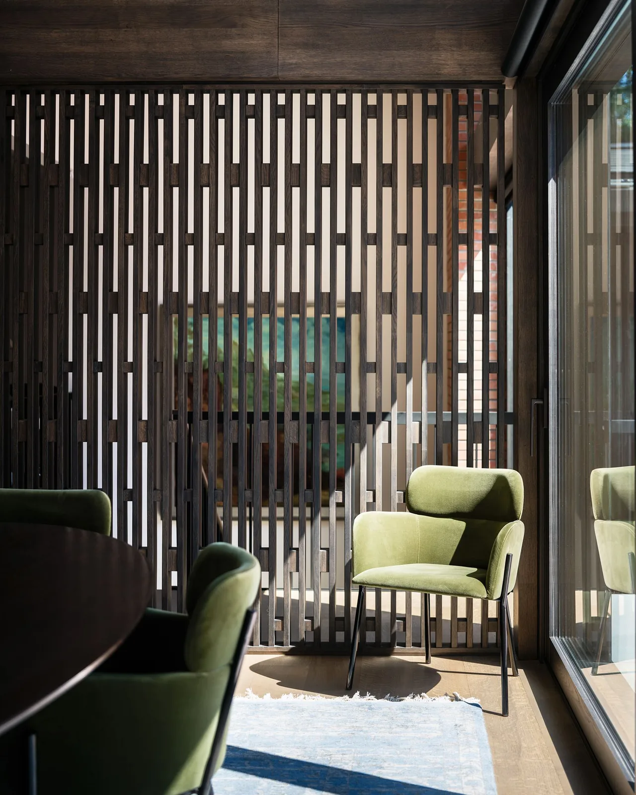 Green velvet chairs sit near a patterned wood slat divider wall in a daylight-filled room with glass doors.