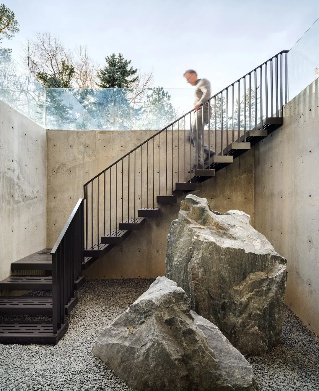 Metal staircase with railing in a sunken concrete courtyard with large rocks and clear glass railing above.