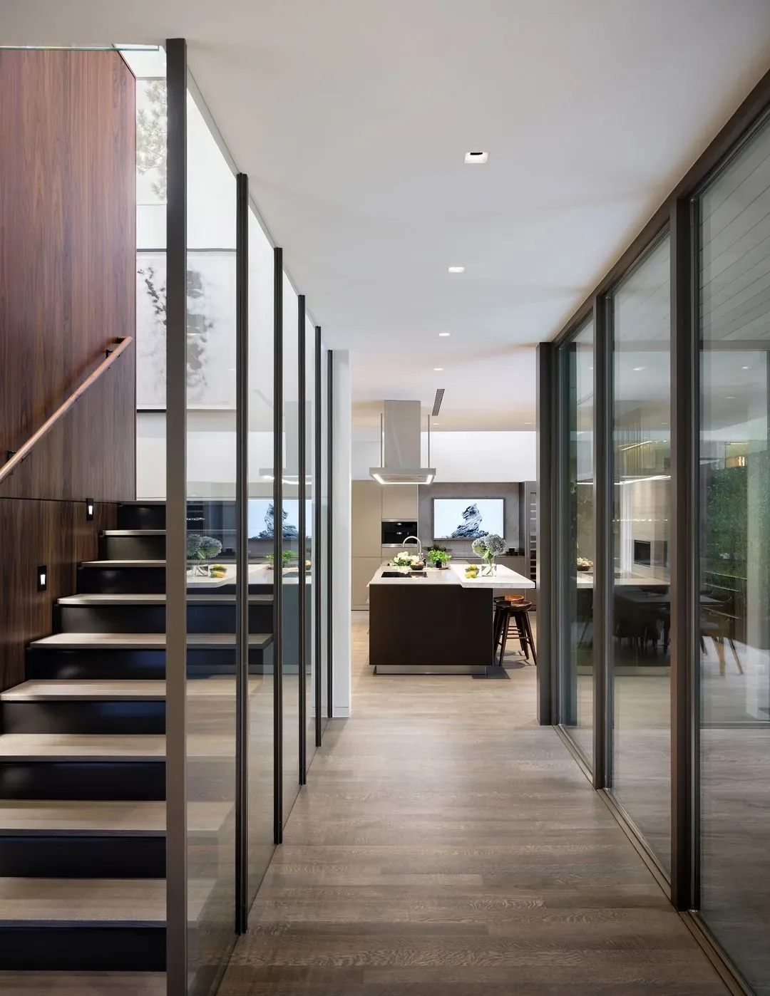 Interior view through a hallway with glass walls and staircase, opening to a kitchen with an island and modern appliances.