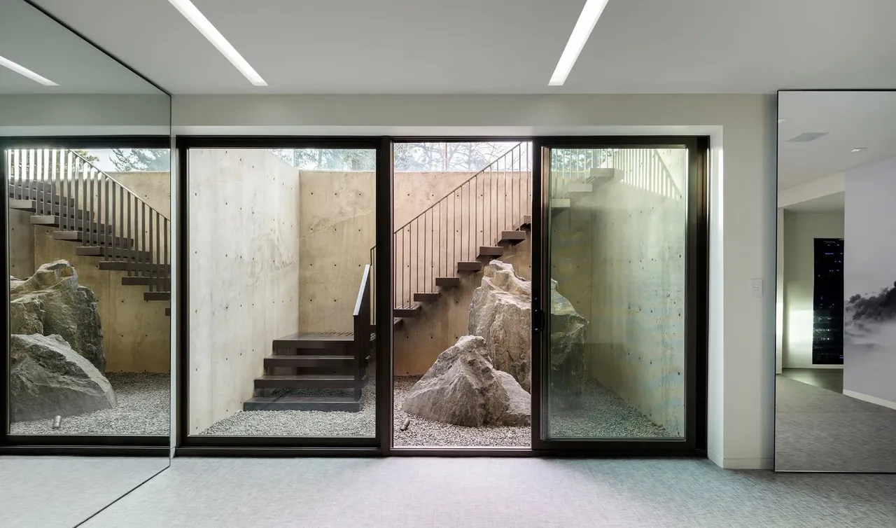 Sunken exterior courtyard with concrete walls, gravel, large natural boulders, and metal staircase viewed through floor-to-ceiling glass doors.