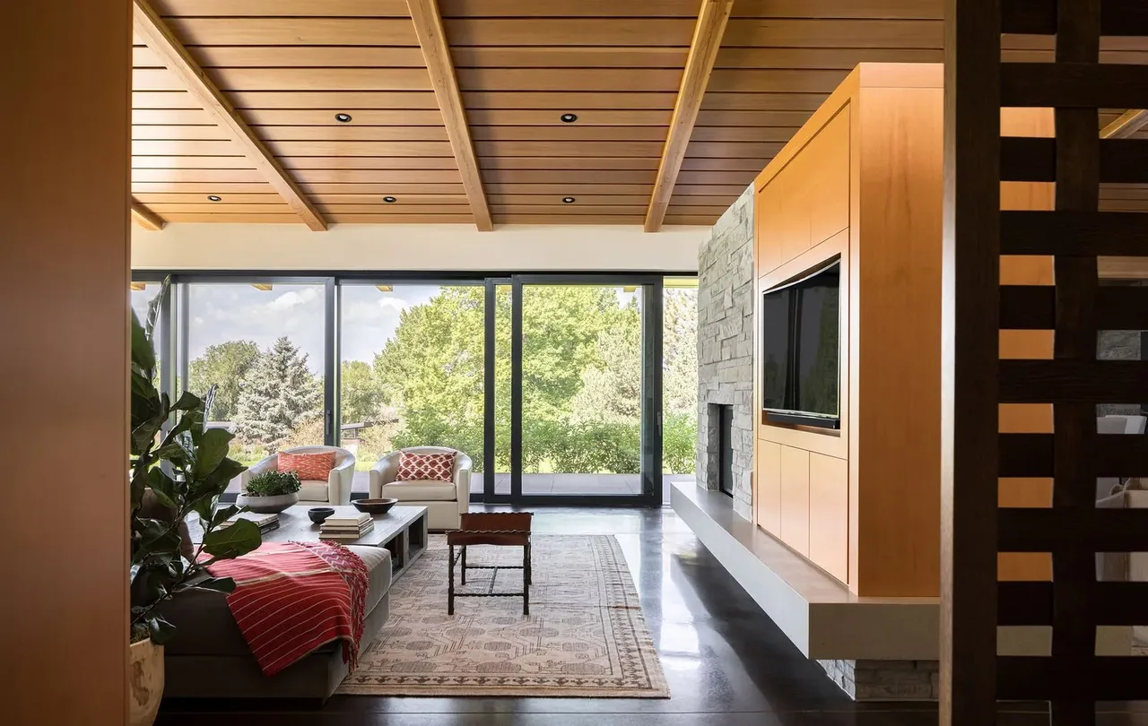 Living area in Cherry Residence features wood ceiling beams, a stone fireplace, and floor-to-ceiling glazing with garden views.