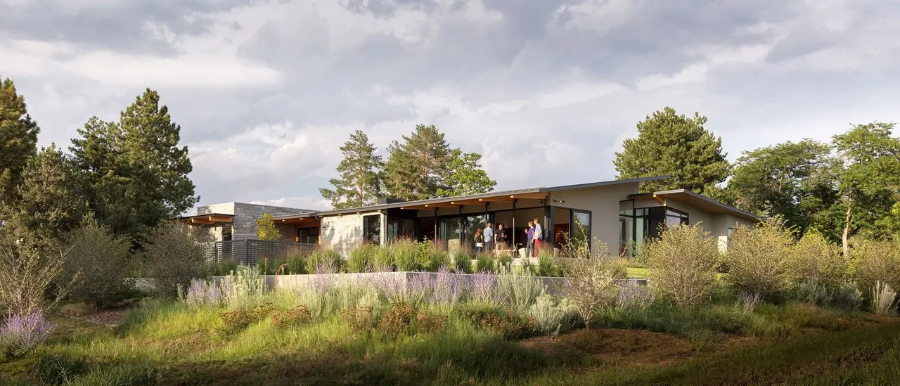 Single-story modern house with large windows, stone and wood exterior, surrounded by trees and native landscaping under a cloudy sky.