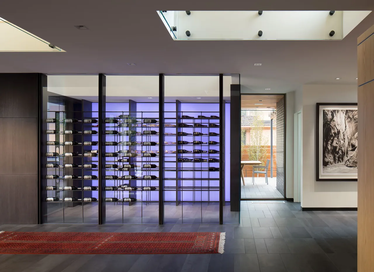 The Duncan Residence hallway features glass-enclosed wine storage with a glowing back panel and a red area runner on dark tile.