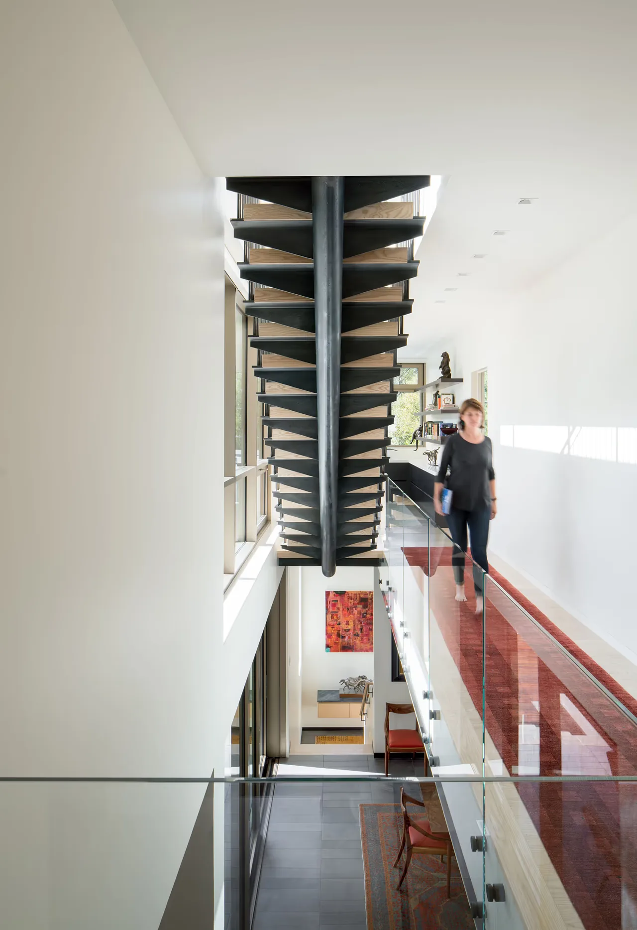 A central black metal staircase is flanked by a glass railing and a red carpet runner in a bright white hallway.