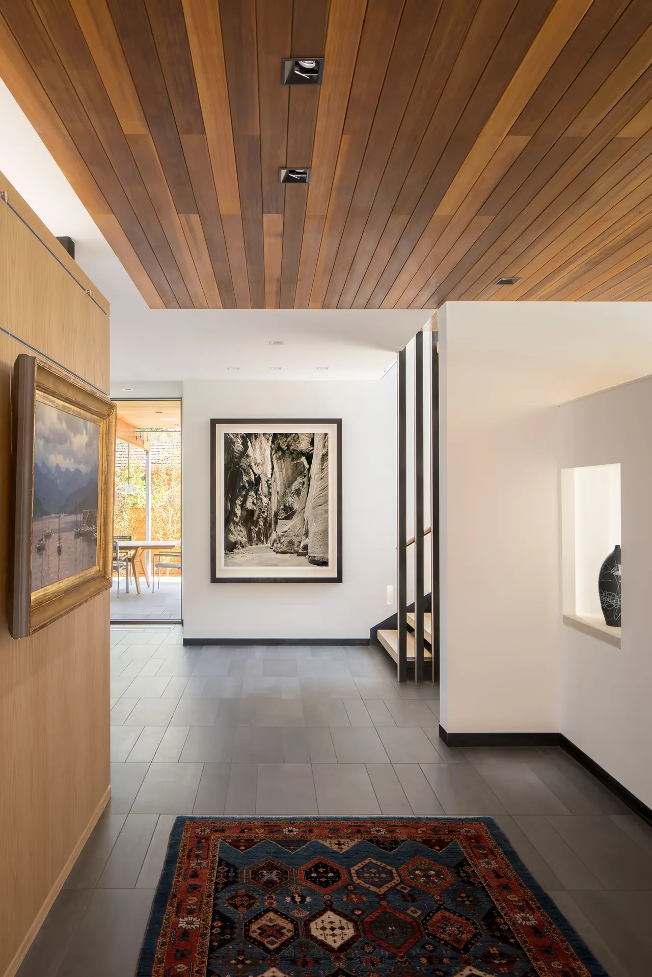 Entry hallway with wood-paneled ceiling and gray tile floor, featuring framed artwork and a decorative rug.