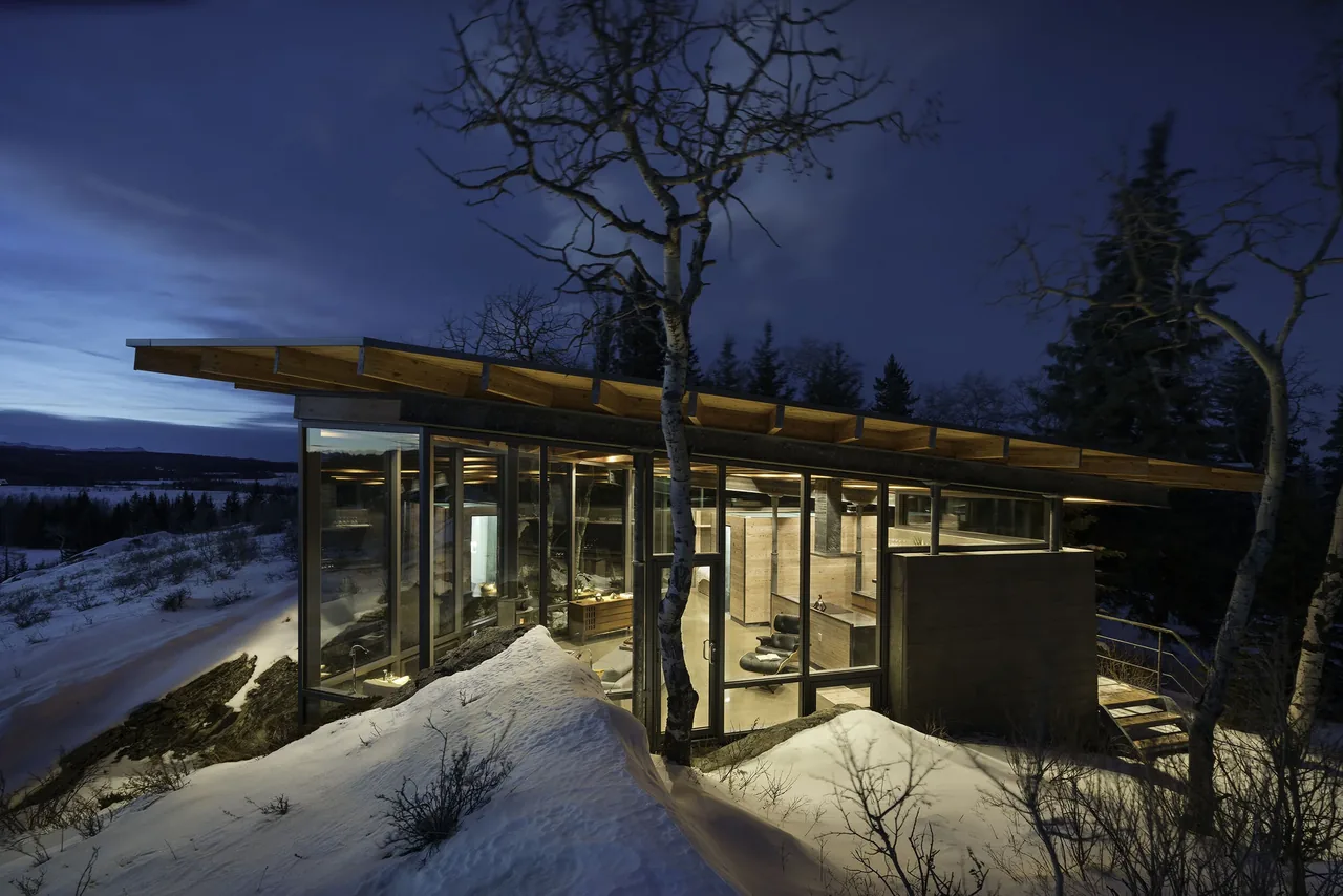 Night exterior front view of ROCK HOUSE featuring floor-to-ceiling glass windows and a flat roof with exposed wooden beams.