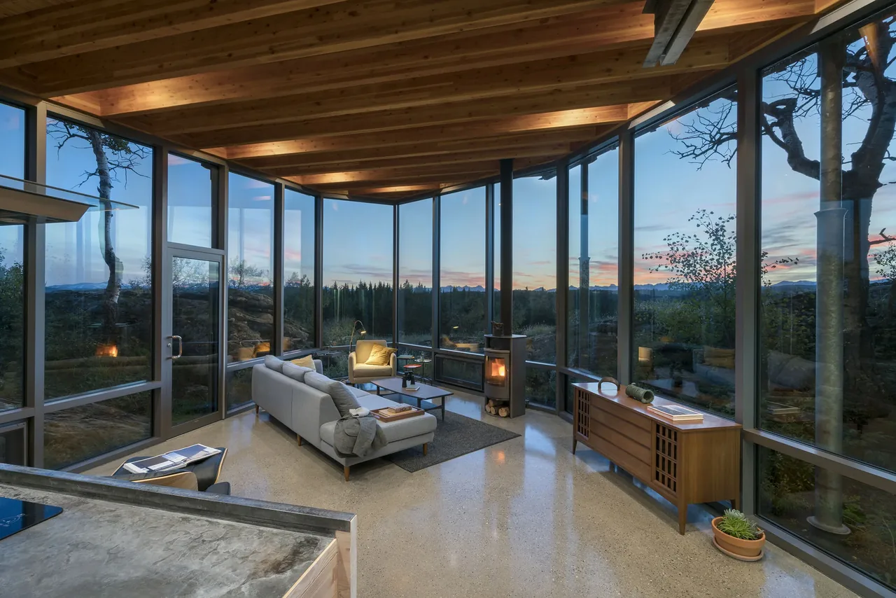 Living room with floor-to-ceiling glass walls and wooden ceiling beams overlooking outdoor scenery at sunset.