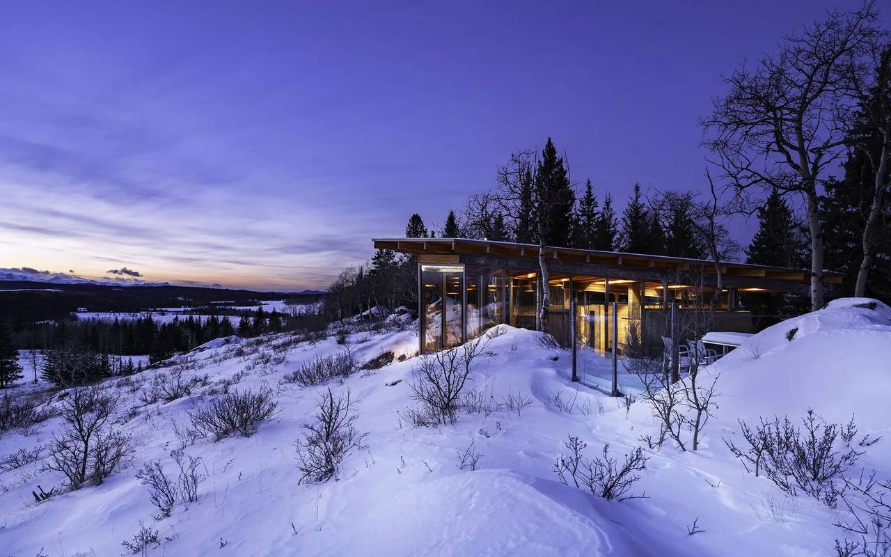 Exterior front view of a modern flat-roofed house with floor-to-ceiling glazing set in a snowy natural landscape at dusk.