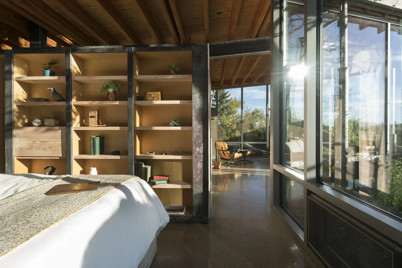 Interior view showing a bedroom with built-in wooden shelves and large floor-to-ceiling glass windows opening to a living area.