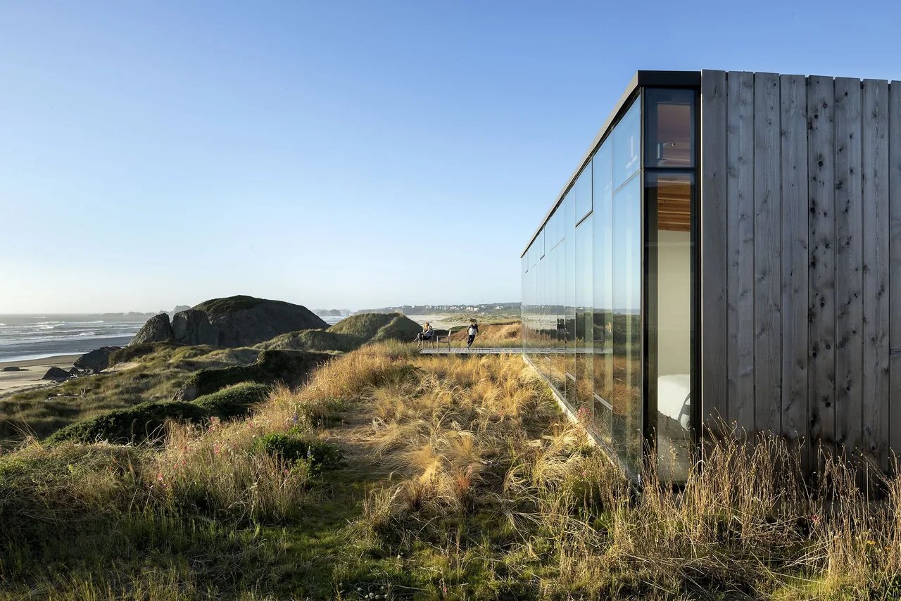 Rectangular beach house with glass facade and wood siding on grassy dunes overlooking the ocean.