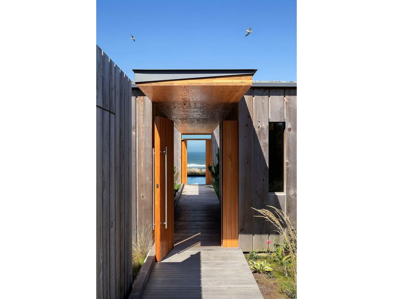 Entry walkway of a beach house with vertical wood siding, a pivot wood door, and ocean view through the passage.