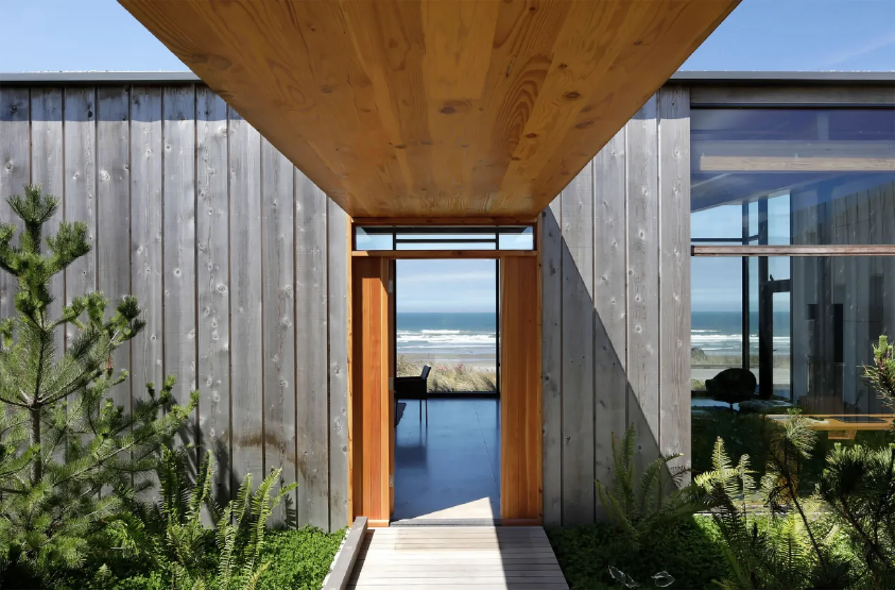 The entrance of the Oregon Coast Beach House features weathered wood siding framed by a wooden ceiling and leads to a view of the ocean through an open door.