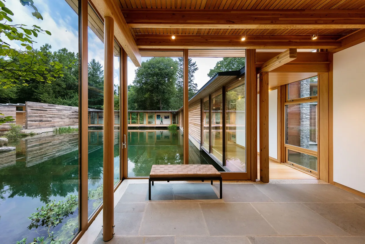 Modern interior space with floor-to-ceiling glass walls facing a pond surrounded by wooden architecture.