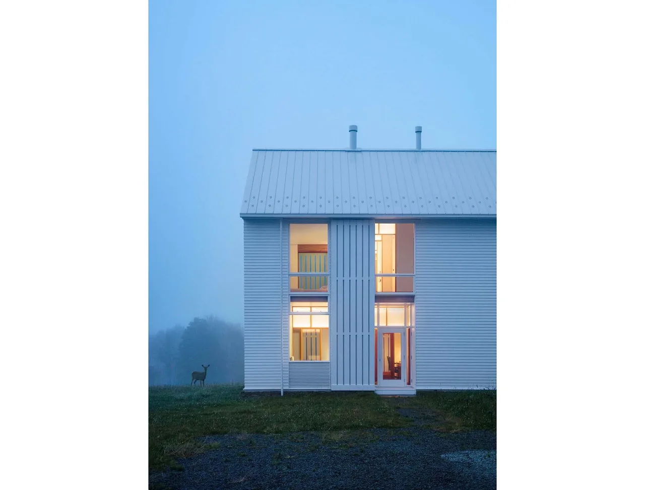 Exterior front view of a white two-story farmhouse with a metal gable roof illuminated during dusk.