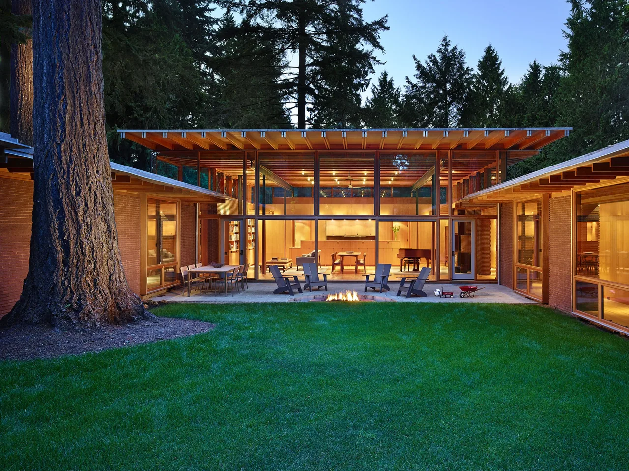 The Beaux Arts Residence features a glass-walled central living space opening to a covered patio with a fire pit, surrounded by brick wings.