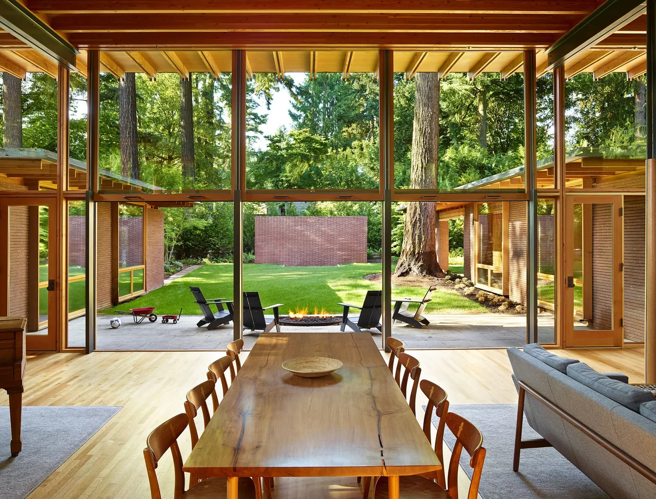 Dining area features a wooden table and chairs with floor-to-ceiling glazing overlooking a backyard fire pit and patio.
