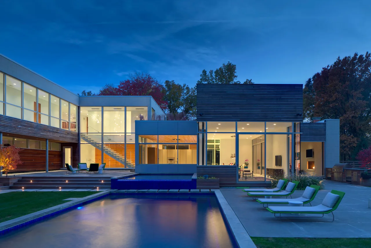 Modern residence exterior at dusk with large windows, deck with lounge chairs, and illuminated swimming pool in the foreground.