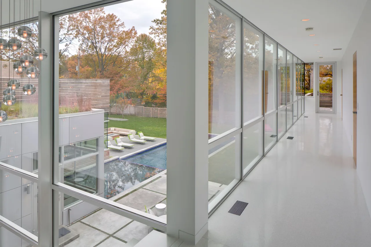 A hallway with floor-to-ceiling glazing provides views of the backyard pool area and lawn.